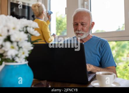 Senior man working on laptop at dining table at home while woman cleaning windows in background Stock Photo