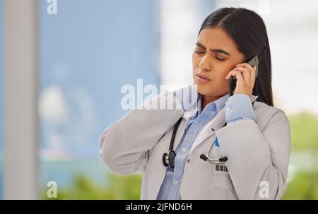 Stressed doctor talking on cellphone in a clinic. Indian healthcare professional standing alone and suffering from neck pain on a busy shift. Medical Stock Photo