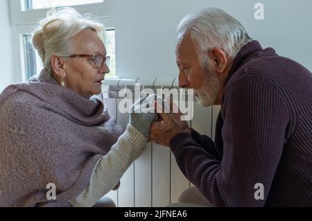 Senior couple holding each other hands beside heater in living room and trying to warm in winter season and energy crisis Stock Photo