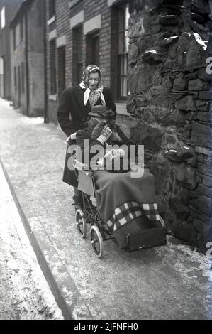 1952, historical, wintertime and well-wrapped up against the cold, an eiderly lady sitting in an old small-wheeled pushchair, with a rug over here, being pushed by a lady in a coat and scarf, along a snow-dusted pavement in old victorian street, England, UK. Stock Photo