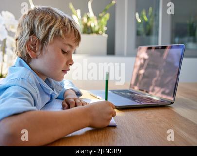 Young homeschool little boy sitting alone and using laptop to study. Caucasian child writing and learning remotely due to covid pandemic. Kid studying Stock Photo