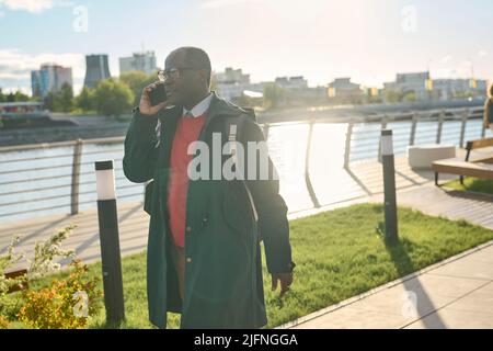 African mature businessman talking on mobile phone while walking along the urban street during sunny day Stock Photo