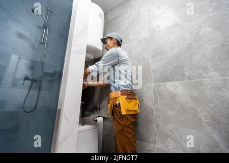Plumber installing electric heating boiler. Young handyman in uniform setting up electric heating boiler at home Stock Photo