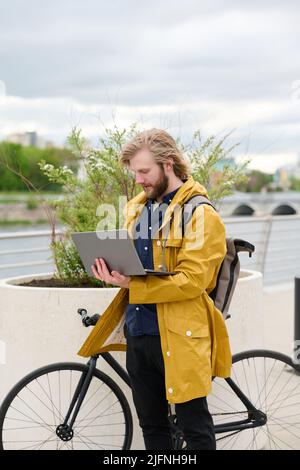 Young businessman using laptop for online work while standing on the street with his bike Stock Photo