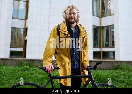 Portrait of young office worker in wireless headphones standing with bike outdoors and smiling at camera Stock Photo