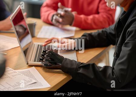 Close-up of young woman with prosthesis arm sitting at table and typing on laptop during meeting at office Stock Photo