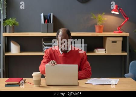 African serious manager in eyeglasses concentrating on his online work on laptop sitting at his workplace at office Stock Photo