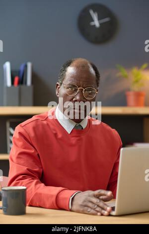 Portrait of African mature businessman in eyeglasses sitting at his workplace with laptop and looking at camera with serious expression Stock Photo