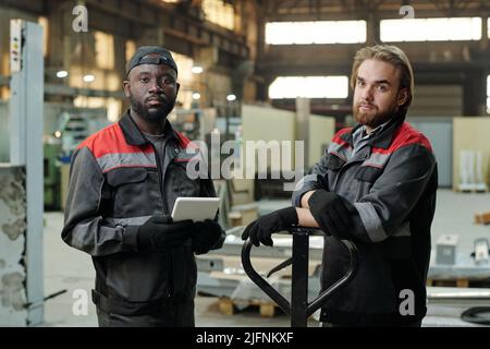 Small group of young interracial male workers of spacious warehouse in workwear standing in front of camera against workshop interior Stock Photo