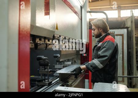 Side view of young engineer in workwear putting metallic sheet on part of working industrial machine or other equipment in factory Stock Photo