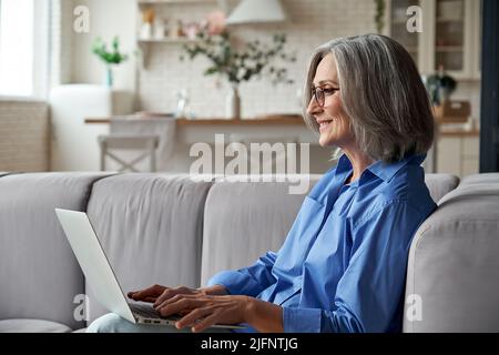 Happy 60s mature woman using laptop computer sitting on couch at home. Stock Photo