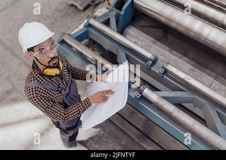 High angle view of heavy industry worker with safety headphones and hard hat in industrial factory holding blueprints Stock Photo