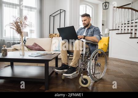 Young male wheelchair user in casual clothes looking at laptop screen during online work in modern living room. Concept of technology and remote job for people with disabilities. Stock Photo