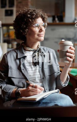 Young pensive businesswoman or freelancer with pen over page of copybook making notes while having coffee and writing down plan Stock Photo