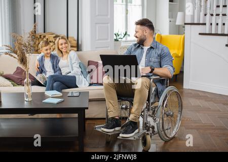 Lovely wife sitting with son on comfy sofa and sincerely smiling while looking at disabled husband. Man in wheelchair working on wireless laptop and spending time with friendly family. Stock Photo
