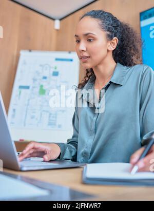 Young happy mixed race businesswoman with curly hair writing in a notebook and working on a laptop sitting at a desk in an office at work. One Stock Photo
