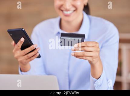Unrecognizable mixed race businesswoman smiling while using a phone and credit card sitting alone in an office at work. Happy hispanic female shopping Stock Photo