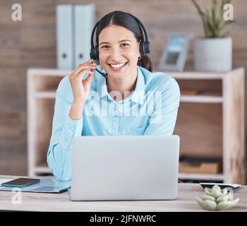 Portrait of a happy smiling hispanic woman wearing a headset and using a laptop while sitting at a desk at her office job. Cheerful young mixed race Stock Photo