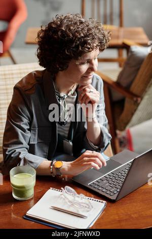 Happy middle aged businesswoman or freelancer networking by table in cafe while keeping hand on chin and looking at data on screen Stock Photo