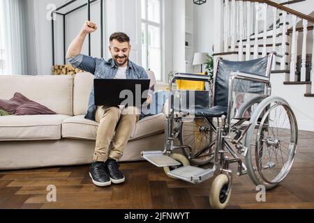 Disabled male sitting on comfy sofa near wheelchair with laptop on knees and doing winner gesture with clenched fist. Happy man winning online lottery and celebrating amazing news at home. Stock Photo