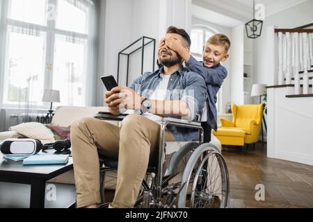 Happy son unexpectedly closing father's eyes from back in modern living room. School boy surprising disabled dad in wheelchair with smartphone in hands. Stock Photo
