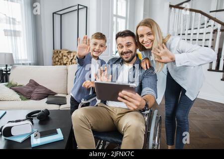 Smiling wife and son standing near disabled father with digital tablet and waving hello while looking at camera. Concept of friendly family and modern leisure entertainment. Stock Photo
