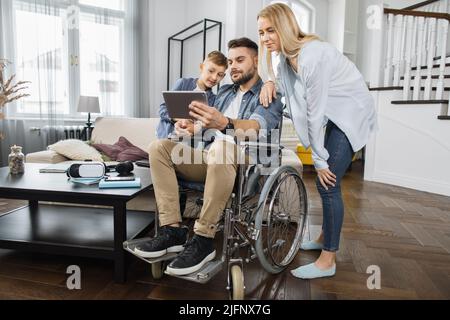 Mother and son looking on tablet screen during video conference while standing near father in wheelchair. Friendly family using modern application for remote communication with relatives. Stock Photo