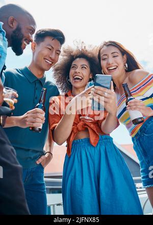 A group of diverse young cheerful friends standing outside together and using a cellphone for selfies and social media. Smiling men and women being Stock Photo