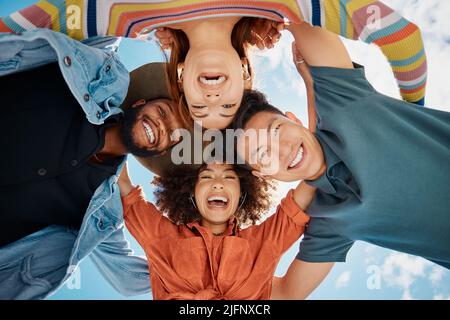 Group of diverse friends standing in a huddle against a blue sky. Multi-racial friends standing together arms around each other and smiling while Stock Photo