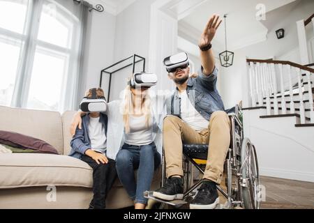 Happy caucasian family with disabled father in wheelchair playing games together using VR headsets. Modern technology for entertainment at home. Stock Photo