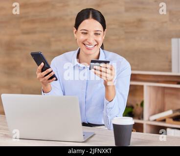 Beautiful young mixed race woman reading her credit card while using her phone and laptop to shop online while sitting in the office at work. Shopping Stock Photo