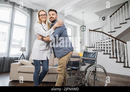 Portrait of positive female physiotherapist and bearded man during rehabilitation at home. Attractive woman embracing disabled man while smiling and looking at camera. Stock Photo