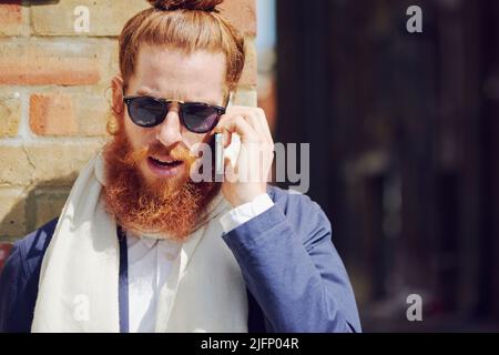 One trendy young fashionable man with man bun wearing sunglasses and talking on a cellphone while standing outside enjoying the sun in the city Stock Photo