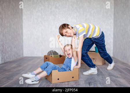 Brother rides sister in a cardboard box. Children play with boxes at home in quarantine. A boy and a girl have moved into a new apartment and are Stock Photo