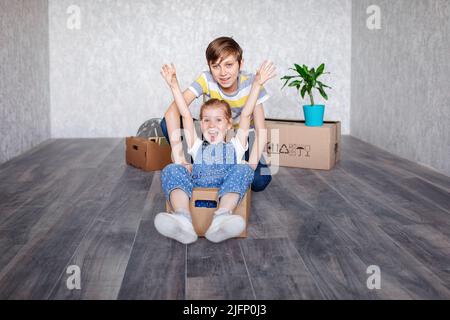 Brother rides sister in a cardboard box. Children play with boxes at home in quarantine. A boy and a girl have moved into a new apartment and are Stock Photo