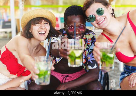Multiethnic group of three people, one African male and two caucasian women having fun drinking cocktails and celebrating toasting with fresh iced coc Stock Photo