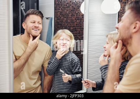Portrait of happy father with cute son looking in mirror in bathroom during morning routine Stock Photo