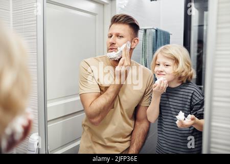 Portrait of father and son shaving together looking in mirror during morning routine in bathroom Stock Photo