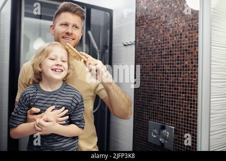 Portrait of happy father brushing hair of little boy and looking in mirror, copy space Stock Photo