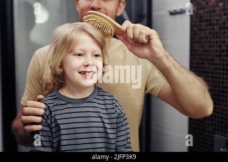 Waist up portrait of blonde young boy looking in mirror with caring father brushing his hair Stock Photo