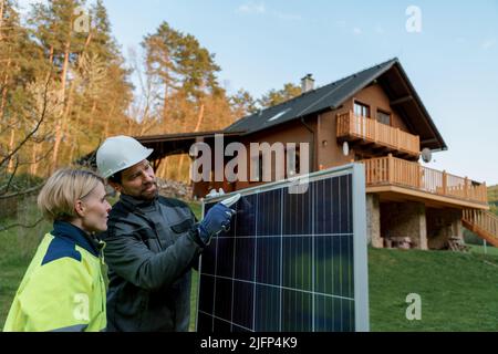Man and woman solar installers carrying solar module while installing solar panel system on house. Stock Photo