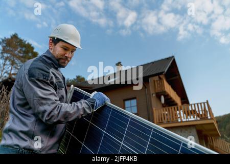 Smiling handyman solar installer carrying solar module while installing solar panel system on house. Stock Photo