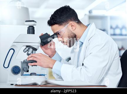 One serious young male medical scientist sitting at a desk and using a microscope to examine and analyse test samples on slides in a hospital Stock Photo