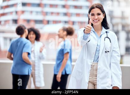 Portrait of a happy young doctor gesturing thumbs up with her colleagues in the background. Group of doctors and nurses collaborating in meeting Stock Photo