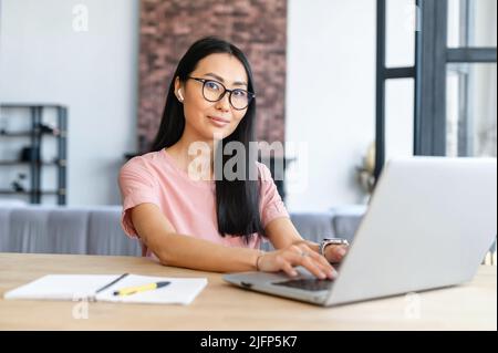 Elegant smart pretty Asian student or entrepreneur in glasses and earphones talking to business partner on the video call and typing, taking notes, and attentively listening and looking at the camera Stock Photo
