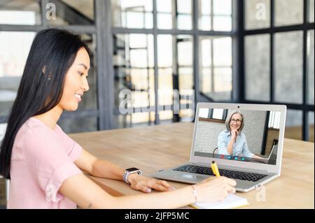 Young Asian woman using application for distance video communication with English teacher, tutor, talking with boss online via laptop, taking a virtual course, studying from home and taking notes Stock Photo