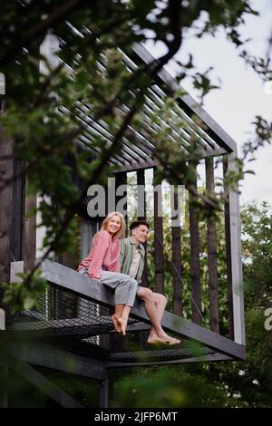 Young couple sitting and cuddling in hammock terrace in their new home in tiny house in woods, sustainable living concept. Stock Photo