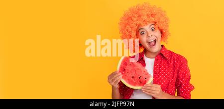 surprised kid in fancy orange hair wig holding fresh ripe water melon slice fruit. Summer girl portrait with watermelon, horizontal poster. Banner Stock Photo