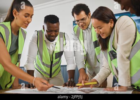 A happy diverse multiracial group of architect colleagues analyzing a blueprint design on a building and discussing plans for the next project. Team Stock Photo