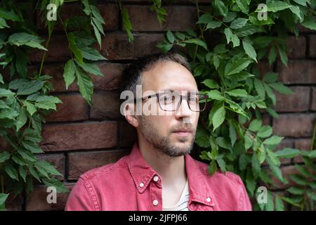 Headshot portrait of smiling 30s caucasian millennial man in glasses sit at street on brick wall Stock Photo